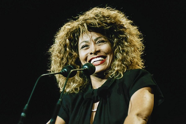 Tina Turner souriante habillée de noir devant deux micros